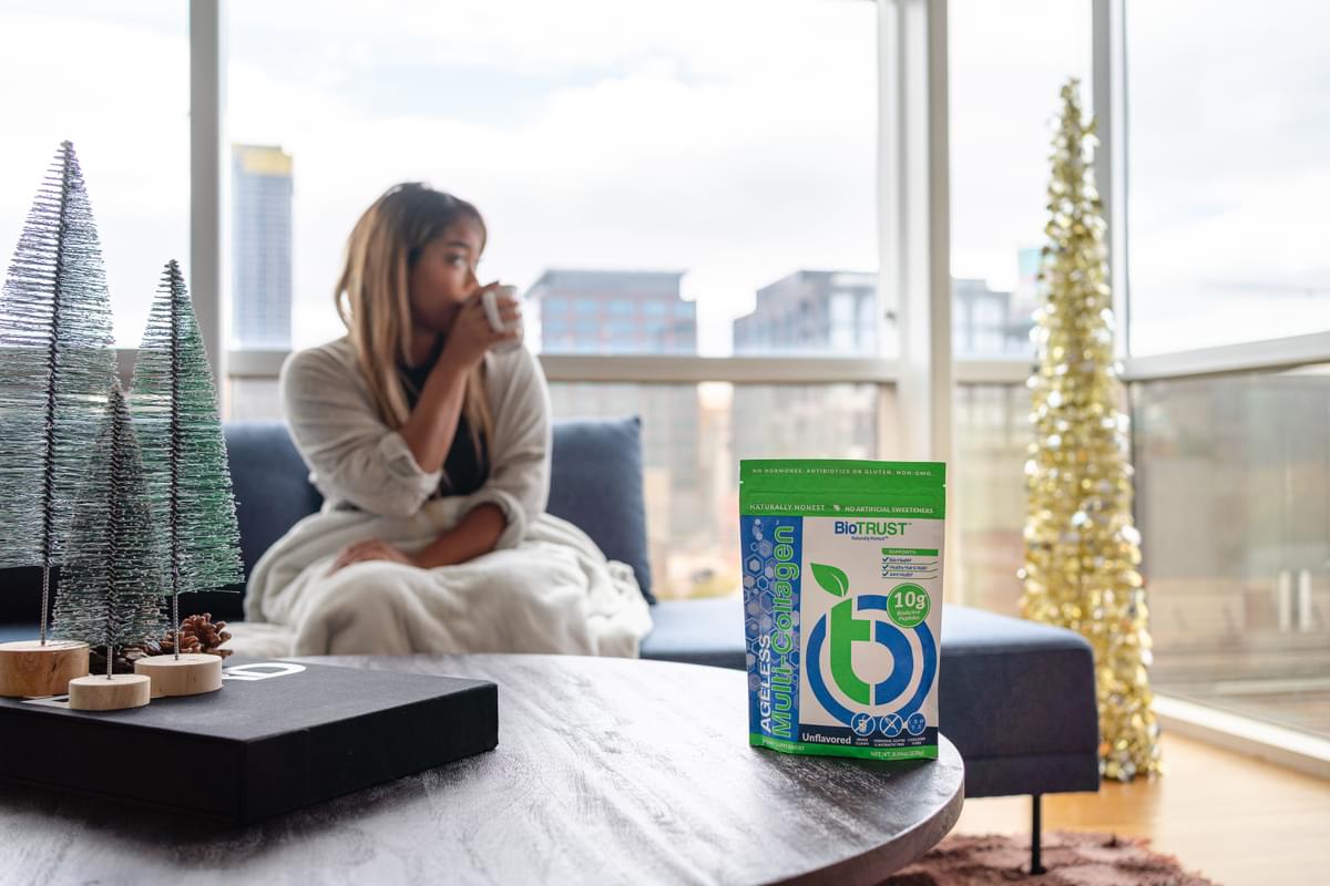 BioTrust Ageless Multi-Collagen pouch on a living-room table with a person enjoying a warm drink in the background